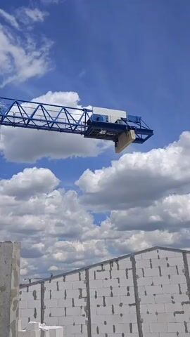 The media file shows a construction scene under a bright blue sky filled with fluffy white clouds. A large blue tower crane dominates the frame, its metallic structure reaching upwards. The crane has a spacious operator's cab with windows, offering a glimpse of a person inside, likely the crane operator. A long, extending boom arm is suspended from the top of the crane, with cables and equipment visible.

In the foreground, construction materials are laid out. On the left, a large, rough concrete slab stands vertically, forming part of a wall. To its right, a wall made of white, rectangular blocks is partially constructed, showing the building process. The overall scene suggests an active construction site, possibly in an open area given the expansive sky and distant industrial-looking structures with smokestacks visible on the horizon.

The lighting suggests it is daytime, likely mid-morning or afternoon, given the brightness and the shadows. The weather appears to be clear and pleasant, with the clouds adding visual texture to the sky. The setting appears to be rural or on the outskirts of a town, judging by the open space and the type of construction. There is no visible text in the media file. The overall mood is one of industrial activity and ongoing development.