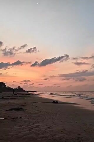 The video shows a sandy beach during a sunset. The sky is a gradient of light blue fading into orange and pink, with scattered clouds. A crescent moon is visible high in the sky. The beach is wide and dark, with the gentle waves of the ocean lapping at the shore. In the distance, silhouettes of trees and possibly people can be seen along the shoreline.  The camera is static.