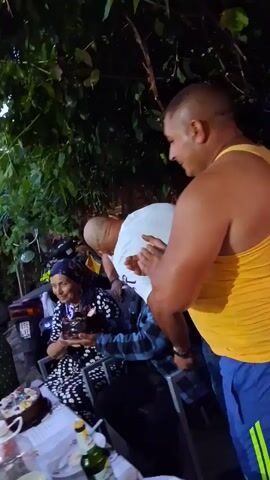 A group of people are gathered around a table outdoors. Some individuals are clapping and smiling. One person, a large man wearing a yellow tank top and blue shorts, is holding a baby. Another person, a woman wearing a headscarf, is seated and appears to be interacting with the baby. The environment suggests a casual gathering, possibly a celebration, with foliage and what appears to be a motorcycle in the background. Bottles and other items are visible on the table. The lighting indicates it is either dawn or dusk.