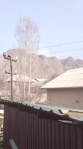 The video depicts a rural landscape in what appears to be early spring. In the foreground, there's a rusty corrugated metal fence. A utility pole with electrical insulators is visible behind the fence, with a bird perched on top. Beyond the fence and utility pole are several buildings with corrugated metal roofs, typical of rural or suburban dwellings. In the middle ground, there is a budding tree with small pink flowers amidst leafless trees and bushes, suggesting the start of spring. In the background, a range of rugged mountains is visible, partially obscured by a light haze. The sky is a pale blue, indicating a clear day. The overall scene suggests a quiet, undeveloped area with a hint of spring growth amidst an otherwise barren winter landscape.