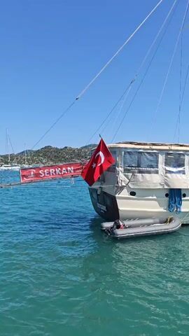 The video captures a scenic boat trip on a bright, sunny day in Demre, Turkey. The main subject is a large, white, two-masted gulet-style boat named "BEYZADE," which is actively cruising on calm, clear turquoise water.

At the stern of the boat, a Turkish flag is flying, and an inflatable dinghy is tethered alongside. Blue ropes are visible hanging from the boat's side. A red banner, partially visible in the initial shots, carries the text "ERKAN YACHT."

The surrounding landscape features lush green, hilly islands or coastal areas under a perfectly clear blue sky.

Passengers are seen relaxing on various parts of the boat's deck. Some individuals are lying on cushions, presumably sunbathing or resting, while others stand, observing the beautiful surroundings. One person is shown standing on an upper deck, actively taking a photograph with a mobile phone, capturing the expansive views. The overall atmosphere appears leisurely and enjoyable, consistent with a holiday or recreational excursion.