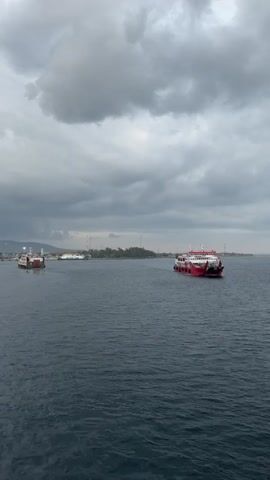 The video begins with a view of the ocean and a cloudy sky. A red and white ferry is visible on the right side of the frame, moving away from the camera. In the distance, a town or port is visible on a strip of land, with several other ferries and boats docked or moving in the water. The camera pans slightly to the left, revealing another ferry. The perspective shifts to show the deck of the ship from which the video is being filmed, with its green deck and yellow and black striped ramp. The video continues to pan left, showing more of the port and the surrounding landscape, which includes mountains in the background. The lighting suggests it is either early morning or late afternoon.