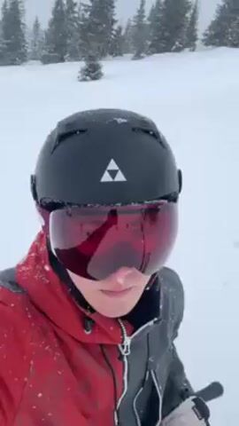 A person wearing a red and black ski jacket, a black helmet with a triangle logo on the front, and red ski goggles is on a ski lift. The person is holding ski poles in their left hand. The ski lift is going uphill over a snowy, tree-covered landscape. Snowflakes are falling. The person sticks their tongue out to catch a snowflake.