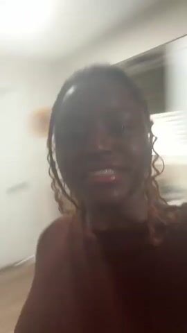 A woman is recording a video in her kitchen. She is wearing a maroon shirt and large hoop earrings. She has brown curly hair, a medium skin tone and glossy lips. She says, "So, we are currently making jollof rice, jollof rice for my..." The camera shifts around so we briefly see a pot on the stove and the kitchen counter.