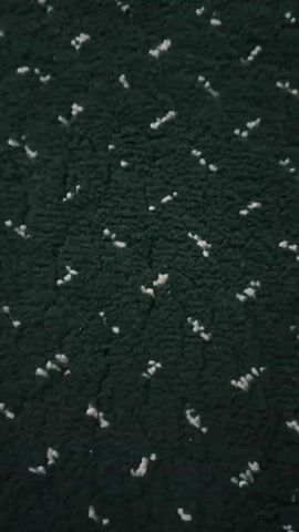 The video shows a close-up of a green carpet with a subtle pattern of white flecks. The camera then pans upwards and to the right, revealing a wider view of a room with more green carpet. This carpet also has a pattern, but it is more distinct, with rows of small white shapes. In the background, there is a person wearing traditional Islamic clothing standing near a wooden cabinet. Further in the background, a wooden door can be seen. The lighting suggests an indoor environment, possibly a mosque or prayer hall, given the carpet design which often signifies prayer rows. The movement of the camera is smooth but slightly shaky.