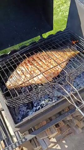 A whole fish is being grilled over hot coals. The fish is placed in a metal grill basket, which is positioned over a black charcoal grill. The fish appears to be cooked to a golden brown color with visible grill marks. Embers and ash are visible beneath the grill basket. The background shows a grassy area and a black grill lid. A pair of metal tongs are visible on the right side of the grill basket. The scene is outdoors, likely in a backyard or park setting.