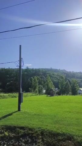 The video shows a rural landscape on a sunny day. The camera pans across a green grassy field with a telephone pole and electrical wires in the foreground. In the distance, a tree-covered hillside rises under a clear blue sky with the sun glinting brightly. As the camera continues to pan, a modern house with large windows and a garage with cars parked outside comes into view. The video appears to be shot from a moving vehicle.