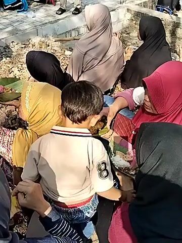 The video shows an outdoor gathering of people, many of whom are seated on the ground. Several individuals are wearing headscarves or hijabs, and some men are wearing black attire and peci hats. There is food being served on banana leaves, and people appear to be eating. In the background, there is a structure with a wooden roof and pillars, and trees are visible. A man holding a camera is standing and filming the event. There are also piles of dirt or soil, and some construction materials or wooden planks are present. The scene suggests a communal meal or a cultural event taking place in a somewhat rustic or natural setting.