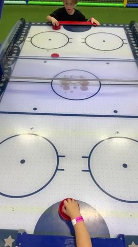Two children are playing an air hockey game in an indoor play area. A young boy with blonde hair, wearing a black t-shirt, stands at one end of the air hockey table, holding a red paddle. A younger child, whose arm is visible in the foreground wearing a wristband, is at the other end of the table, also holding a red paddle. The air hockey table is blue and white with colorful flashing lights along the sides. The game appears to be in progress, with a red puck visible on the table. The background shows a brightly colored soft play area with green turf and yellow and blue netting, consistent with an indoor children's entertainment center. The lighting suggests it is daytime, likely indoors. The children appear engaged and focused on the game. The number "2" is visible on the blue front panel of the air hockey table. No other visible text is present. The setting is likely an amusement or play center in Sovkhoza imeni Lenina, Russia.