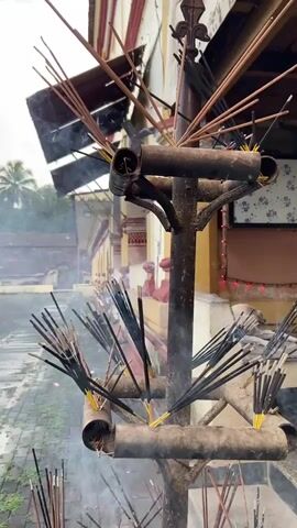 A metal incense stand with multiple sticks burning is in the foreground. There is smoke rising from the incense. In the background, there is a building with yellow and red trim. A dog is sitting on a ledge. Several people are walking in the background.