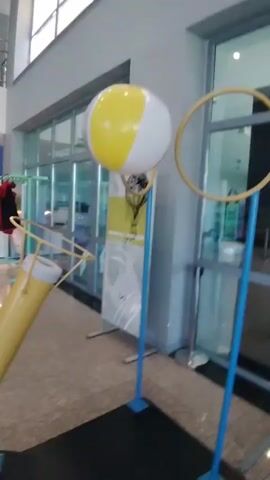 The video features an indoor science exhibit demonstrating Bernoulli's principle. A large yellow cannon-like device, angled upwards, is shown on the left. It generates an upward air current, suspending a large beach ball in the air. Initially, the ball is yellow and white. Later, a different beach ball, colored yellow, green, and white, is seen floating in the same position. The ball remains suspended above the air source, wobbling slightly but generally staying within the vicinity of a yellow circular hoop supported by blue stands to the right. In the background, a poster is visible with the text "BERNOULLI PRENSİBİ," which translates to "Bernoulli's Principle" in Turkish. The setting appears to be a modern building, likely a science museum or exhibition space, characterized by large glass windows, tiled flooring, and bright ambient lighting. No people are in focus, and the scene conveys an educational and observational atmosphere. The exhibit illustrates how moving air can create lift and stabilize an object.