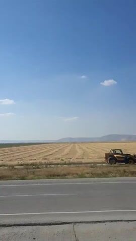 The video shows a landscape with a harvested field and a road in the foreground. A yellow tractor with a loader attachment is visible in the field. The field has rows of cut crop, likely wheat or barley, lying on the ground. The sky is blue with a few scattered clouds. In the distance, there are hills or mountains. The road appears to be asphalt and there is a curb or edge on the side of the road. The overall scene suggests a rural agricultural setting during harvest time.