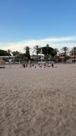 A group of approximately ten people are practicing yoga on a sandy beach. They are arranged in a circle on white yoga mats. The people appear to be a mix of adults and children. The background shows palm trees, buildings, and a clear blue sky. The tide is out, exposing a wide expanse of sand. The sun appears to be setting, casting a warm glow over the scene.