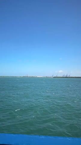 A ferry, viewed from its deck, travels across a body of water under a clear blue sky. The water is a turquoise green and shows moderate waves with white caps. In the distance, a long coastline is visible with a line of trees and buildings, including several large industrial cranes. A large cargo ship is visible on the water. Part of the ferry's railing, painted blue, is in the foreground. The sky is mostly clear with a few scattered white clouds.