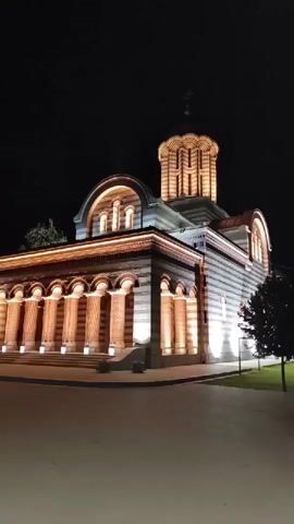 The video shows a nighttime view of a building with an arched entrance and a domed tower. The building is illuminated with warm lights, highlighting its architectural details, including columns, arches, and striped facade. A tree is visible to the right of the building. The ground in front appears to be paved. The sky is dark, indicating it is night.