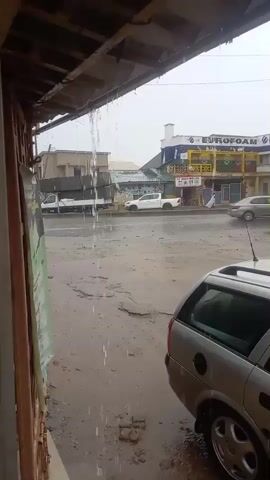 The scene shows a rain-soaked street in what appears to be a developing country. Rain is falling heavily, with water dripping from a corrugated roof in the foreground. Across the street, a building with the sign "EUROFOAM" can be seen, along with other storefronts. Cars and a three-wheeled vehicle traverse the street. The road surface is wet and patchy. A silver station wagon is parked to the right of the frame, partially obscuring the view.