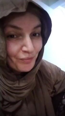 The video is a close-up of a woman's face as she eats a piece of chicken. She is wearing a headscarf and a brown top. The background is out of focus. The woman is holding the chicken with her right hand and bringing it to her mouth. She appears to be enjoying her meal.