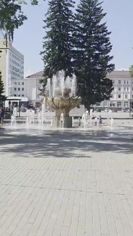 A vertical video opens on a sunny day in a plaza with a large, ornate fountain in the center. The fountain has multiple tiers and jets of water spraying upwards. Several smaller fountains are located around the main one, creating a watery play area. In the background, there are buildings, trees, and some people. The ground is paved with light-colored interlocking bricks. Two tall evergreen trees stand behind the fountain, framing it. In the distance, a multi-story white building with many windows is visible on the left. To the right of the fountain, there are more buildings, including a light-colored one with several windows and a darker tiled roof. People are visible near the fountain, with some appearing to be children playing in the water. The overall scene is one of a public space on a bright, warm day, with a focus on the fountain and the people interacting with it.