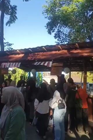 The video shows a crowded outdoor area beneath a covered structure with a thatched roof. A group of people, many wearing hijabs, are gathered. Some are standing, while others are seated on benches. The atmosphere appears to be lively, with people interacting and moving around. The environment is characterized by trees and a clear blue sky. The lighting suggests it is daytime, possibly late morning or afternoon, given the brightness and the shadows. There are decorative elements, such as striped banners, hanging from the roof. Some individuals are carrying bags or backpacks. The people are dressed in casual clothing. The camera is positioned to capture the scene from a slightly elevated viewpoint, looking towards the gathering.