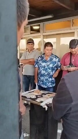 A group of four men stand around a small black table with a game laid out on top of it. One of the men bends over the table, manipulating something with his hands, while the other three watch. The table is cluttered with cards and what appears to be money. The man manipulating the objects at the table is wearing a black shirt and has sunglasses tucked into the back of his head. The man to his left wears a blue Hawaiian shirt. The man behind him has his arms crossed and is wearing a tan baseball cap and polo shirt. The man to the right is wearing a pink shirt and black baseball cap. The men are in front of a white building with windows. A pole is partly visible in the foreground on the left side of the frame.