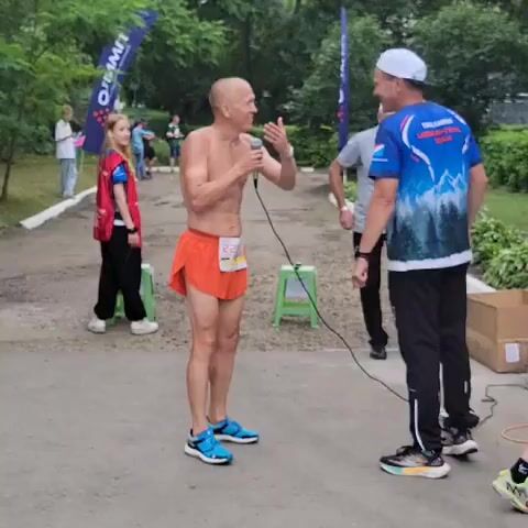 A man with a shaved head, wearing orange shorts and blue running shoes, is speaking into a microphone. He is shirtless and appears to be at an outdoor event. To his right, another man in a blue t-shirt with a mountain graphic and a white baseball cap faces him. A third man, wearing a grey shirt and dark pants, is bending down to the left of the man with the microphone. In the background, two blue flags with white lettering are visible, along with several other people and trees. A woman in a red vest is also visible in the background. The ground is paved.