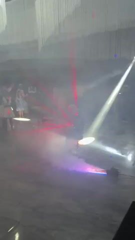 This video captures a lively party scene with flashing red and blue lights and fog effects. Several people are visible in the background, appearing to be dancing and socializing. The focus of the video is a person standing in the center of the room, illuminated by red laser beams that create a dynamic light show. A fog machine is actively producing a thick cloud of smoke, which enhances the visual effects of the lights. The floor is tiled, and the overall atmosphere suggests a celebratory event like a wedding reception or a club night.