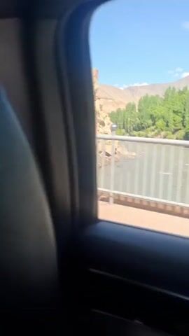 The video is taken from inside a moving vehicle, likely a car, traveling across a bridge. The camera focuses on the view from the passenger window. Outside, there's a landscape featuring a river or wide stream, flanked by greenery on one side and rocky, arid terrain on the other. The river is visible between the bridge's metal railings.  In the distance, there are mountains under a clear blue sky. The vehicle is in motion, as the scenery shifts slightly.