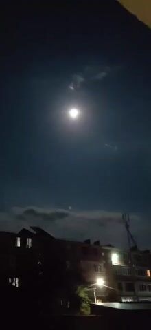 The video shows a nighttime view of a dark blue sky with a bright full moon. The moon is surrounded by a soft glow and a few wispy clouds are visible. Below the sky, the silhouetted roofline of several buildings is visible. Some windows in the buildings are lit, suggesting occupancy. A tall antenna or tower is also visible on the right side of the frame. The overall mood is serene and quiet.