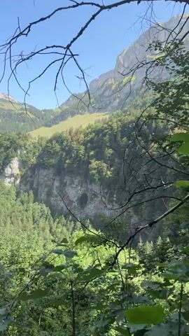 The video pans across a scenic view of a lush green valley with steep, rocky cliffs on either side. A dense forest covers the valley floor and climbs the slopes of the cliffs. In the distance, majestic mountains with rugged peaks rise against a clear blue sky. The camera moves to reveal a path in the foreground where a small group of people can be seen walking. The overall scene depicts a beautiful natural landscape with a hiking trail.
