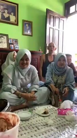 The video shows a group of people gathered in a room, sitting on the floor around a spread of food. Several women wear hijabs, and some are dressed in light blue outfits. A man with a shaved head and a sleeveless shirt sits among them. In the center of the group is a large white bucket filled with what appears to be a mix of chocolate and strawberry ice cream. People are eating the ice cream from small cups with spoons. There are also bottles of red liquid, small plates with food, and tissues scattered on the floor. Some individuals are talking and laughing, while others are using their phones. The room has green walls adorned with framed pictures, and a wooden cabinet is visible in the background. A partially open door reveals some outdoor light.