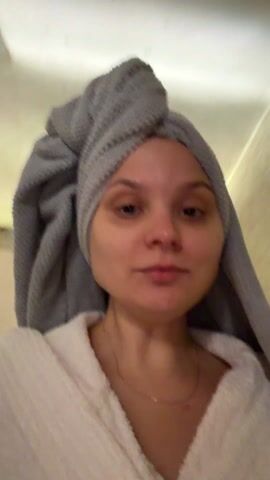 A young woman is featured in a close-up, slightly shaky video, suggesting she is holding the camera herself. She is indoors, possibly in a hallway, with plain, light-colored walls visible in the background. The lighting is somewhat dim.

She is wearing a white bathrobe and has a grey towel wrapped around her head, indicating she has recently showered or is preparing for a bath/shower routine. Her face is visible and appears to be without makeup. Her eyes look a bit tired, with slight puffiness under them. She has light-colored hair, mostly covered by the towel. A delicate gold chain is visible around her neck.

The woman is speaking, and her mouth is moving as she utters the words "All day, all night. What the f***." Her expression is relatively neutral, though the words suggest a feeling of weariness or exasperation. She moves slightly during the video, which contributes to the mild camera movement.