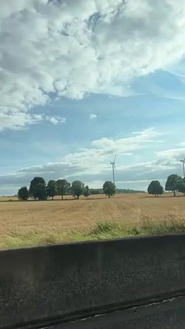 This video captures a view from a moving vehicle, likely a car, looking out at a rural landscape. The camera pans slightly, showcasing a golden field of harvested crops, possibly wheat or barley, stretching towards a line of trees. A solitary wind turbine stands tall in the distance, a modern element against the natural scenery. The sky is partially cloudy with bright blue patches. The foreground is dominated by a concrete barrier, typical of a highway or major road, and the edge of the road itself is visible. As the video progresses, the perspective shifts slightly, and trees pass by, temporarily obscuring the view of the field and wind turbine. The scene then transitions to a green field, possibly corn, under a sky with more dramatic cloud formations, suggesting a change in weather or location. The overall impression is of a drive through the countryside on a partly cloudy day.