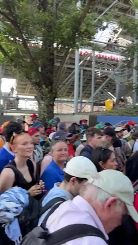 The video captures a bustling scene with a large crowd of people. Many individuals are wearing hats, with a notable presence of green and red caps. Backpacks are also visible on several people. In the background, a large black sign displays a map of a race track, identified as the "FORMULA 1 GRAN PREMIO DE ESPAÑA CATALUNYA." There are also structures resembling grandstands and trees lining the area, suggesting an outdoor event, likely a race. The crowd appears to be moving or gathered around the sign. The lighting suggests daytime, with clear skies.