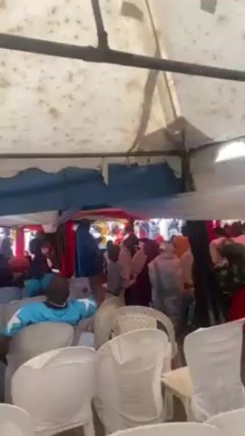 The video shows a crowd of people under a tent, seated in white chairs. The tent is white with a blue trim.  The crowd is dressed in a variety of clothing, including colorful patterns, traditional attire, and casual wear. Several children are visible, including one in the foreground. The ground is not clearly visible, and the lighting is moderate.  Some people are looking towards a focal point, likely a stage or activity area. The tent appears to be set up for an event.