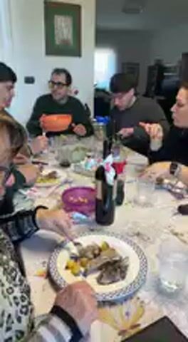 A group of approximately six to seven people, mostly adults, are gathered around a dining table in what appears to be a home setting in Aradeo, Italy. The scene depicts a casual mealtime event.

The individuals are seated at a rectangular table covered with a light-colored tablecloth. Each person has a plate, cutlery (forks and knives), and a glass for beverages. A bottle of red wine and glasses of water are visible on the table. The plates hold servings of food, which include what looks like pieces of meat (possibly pork or lamb), green chopped vegetables (such as celery), and golden-brown fried items, likely potatoes or another vegetable. One person is seen cutting their food, while others are actively eating or reaching for dishes.

A woman with long blonde hair, wearing a light blue top, holds a large, round serving pan filled with the fried potato-like food, suggesting she is either serving or has just served the dish. A man with short hair and a dark shirt is focused on his plate, using a knife and fork. Another man with glasses is visible in the background, also eating. The atmosphere appears relaxed and conversational.

Notable objects include a mobile phone resting on the table, framed artwork on the walls, and typical household decor in the background. The lighting suggests it is daytime, possibly a lunch or early dinner gathering. There is no discernible text visible in the video.