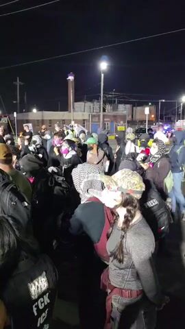 The video captures a nighttime scene in Newark, United States of America, depicting a confrontation between a large crowd and a line of police officers.

The initial shot displays a dense gathering of people, many of whom are wearing face masks (ranging from surgical to fabric) and carrying backpacks. One individual in the crowd wears a black vest with the text "WMB FCK ICE" partially visible, while another wears a "PRESS" vest. The crowd is vocal, with chants such as "We want our streets!" and "We want what we want! What do we want? Justice!" clearly audible. In the background, industrial structures, including a tall chimney, are illuminated by streetlights and overhead wires.

The second shot focuses on a line of uniformed police officers. Their uniforms bear "NEWARK POLICE" patches, confirming their affiliation. One officer's name tag is visible, reading "M. JACOBS". The officers stand in a formation, observing the crowd, with some looking forward and others to the side. A person in a wheelchair is also visible among the police personnel. The setting remains outdoors at night, with streetlights providing illumination, and tents or canopies are discernible in the background. The overall atmosphere appears tense due to the verbal exchanges and the standoff between the two groups.