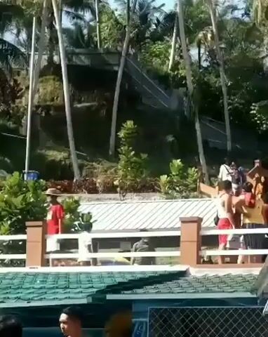 A video shows a natural waterfall cascading down a rocky incline surrounded by lush vegetation and palm trees. In the foreground, several people are visible standing behind a white fence. One person, wearing a red shirt and a hat, is near the center of the frame. To their left, two individuals are wearing headscarves, one in yellow and one in black. The fence itself appears to be made of white posts and horizontal rails. Below the fence, a blue pool of water is partially visible, reflecting the sky. The overall scene suggests an outdoor recreational area or a tourist attraction, possibly a resort with a waterfall feature. The lighting indicates it is daytime with bright sunshine.
