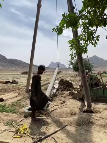 The video captures a daytime scene of a drilling operation in Nowruzi, Afghanistan. In the foreground, a man in dark traditional clothing walks across dry, dusty ground. To his right, green foliage from a tree partially obstructs the view. A large drilling rig stands prominently, featuring two tall vertical poles and a long, suspended pipe or casing. Near the rig, machinery with a prominent green, spoked wheel is in operation, making a loud mechanical sound, likely related to the drilling process. Another individual can be partially seen operating this machinery. The ground is barren and arid, with a discarded tire and metal pipes lying around. In the background, a vast, open landscape with distant mountains under a clear sky completes the scene, suggesting a remote or rural setting. No text is visible in the video.
