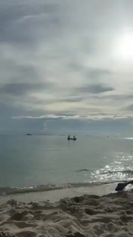 The video shows a tranquil beach scene under a cloudy sky with the sun shining through. The beach consists of light-colored sand, with gentle waves lapping at the shore. In the distance, a small boat is visible on the water with two people standing in it. The water appears calm and clear, reflecting the sunlight. The overall atmosphere of the scene is peaceful.