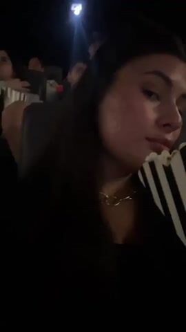 A person takes a selfie at a movie theater. The person is holding a container of popcorn and is surrounded by other people. The environment is dark, with the light from the movie screen illuminating the space. The person has long, dark hair and is wearing a gold chain necklace. The camera is close to the person's face, providing a clear view of their features.