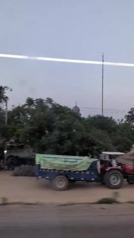 The video captures a rural landscape with a red tractor parked in the foreground on the left. Beyond the tractor, several trees are visible, some with lush green foliage and others appearing sparser. In the background, a domed structure, possibly a temple or religious building, stands prominently against a pale sky. A tall, slender pole is also visible near the domed structure. The ground appears to be unpaved and somewhat uneven, with mounds of dirt or sand in places. A truck with a yellow and brown tarp is seen driving past in the mid-ground. The overall atmosphere suggests an outdoor scene in a somewhat undeveloped or agricultural area.