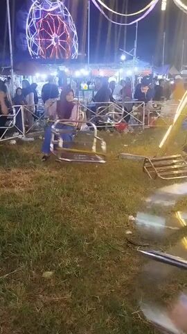 This is a low-angle, handheld video of a carousel ride at night. The ride appears to be a spinning amusement park ride with several seats that rotate around a central axis. In the foreground, several of the shiny metallic seats are blurred due to motion. In the middle ground, two women are visible on the ride. The woman on the left is wearing a blue headscarf and a pink top, while the woman on the right is wearing a purple headscarf and a pink top and blue pants. Both women are in motion, appearing to be enjoying the ride. In the background, a large ferris wheel with colorful lights is visible. There are also several tents and a crowd of people walking around, suggesting this is a fair or festival. The ground is covered in grass.
