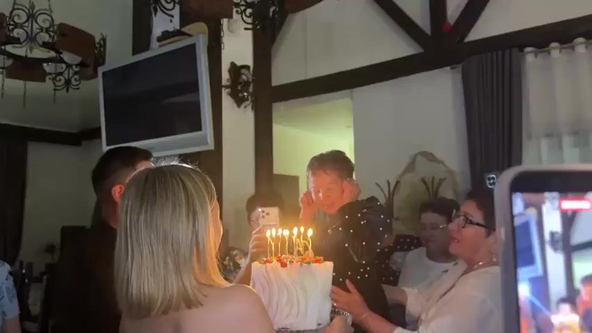 This media file captures a birthday celebration in Zelenaya Polyana, Russia. The scene is indoors, likely a restaurant or a private home, with rustic wooden beams adorning the ceiling. A rectangular television is mounted on the wall, and a chandelier hangs above.

The central focus is a white birthday cake with seven lit candles. An older woman, the birthday honoree, is leaning forward, her hands cupped around her ears, as she blows out the candles. Her eyes are closed, and her mouth is pursed in concentration. A younger woman with blonde hair, seen from behind, stands close to the cake, appearing to be part of the celebration. Several other people, mostly women of varying ages, are gathered around, observing the moment. Some are holding up phones, presumably to record or photograph the event, while others look on with smiles. One woman in the foreground is holding a smartphone, displaying a live video feed of the scene on its screen. The lighting suggests it is nighttime or an indoor setting with artificial light. The overall mood is festive and joyful, with a sense of family or close community evident in the shared experience. No visible text is present in the media.