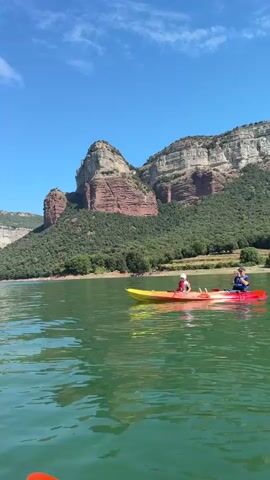 The video opens with a wide shot of a tranquil, green lake under a bright blue sky with scattered clouds. In the foreground, the water is calm with gentle ripples. A two-person kayak is visible in the middle of the frame, with two individuals inside. One person is in the front and the other is in the back, both appearing to be paddling. Behind the kayak, the shoreline is lined with lush green trees that ascend the steep slopes of a massive, multi-layered rock formation. The rock face exhibits distinct horizontal striations of reddish-brown and lighter beige rock, creating a dramatic visual contrast with the verdant foliage.

The scene then transitions to a medium shot of a stone pyramid-like structure partially submerged in the lake. The pyramid is made of rough, pale stones and has a weathered appearance. At its apex, a simple cross-like structure topped with a flag is visible. The water laps at the base of the pyramid, indicating a high water level. The background shows the same rocky mountains and tree-covered slopes as the previous shot, under a clear blue sky.

The video returns to a wider view of the lake and the kayak, showing the two individuals continuing their journey on the water. The kayak, a tandem model, is predominantly yellow and red. The person in the front is wearing a red life vest and a white hat, while the person in the back is wearing a blue life vest. They are both looking forward, seemingly enjoying the scenery. The vast, majestic mountain range serves as a striking backdrop to this serene water activity. The light suggests it is daytime, likely mid-morning or afternoon, given the brightness and clear skies. The overall atmosphere is peaceful and recreational.
