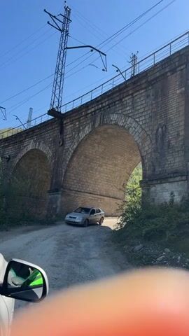 The camera looks out from the driver's side window of a car, looking up at a stone arch bridge. The road is dirt, and there is a parked silver sedan under the center of the bridge. Power lines and a metal tower cross the top of the frame. The sky is blue. Green vegetation is on the ground under the bridge.