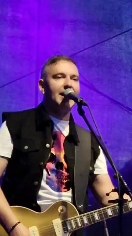 A man in a white t-shirt with a skull graphic and a black vest is playing an electric guitar and singing into a microphone. He has short hair and is wearing an earring in his left ear. He is on a stage with a purple backdrop.