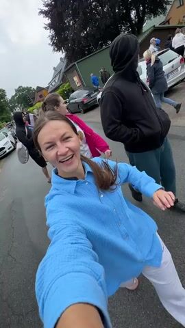 A group of four people, a woman in a blue shirt, a man in a black hoodie, and two women in the back, walk down a street. The woman in the blue shirt is recording a selfie video, smiling at the camera. The man in the black hoodie behind her also smiles and gestures with his hand. The two women in the background are looking at the camera and smiling. The street is lined with parked cars and houses with green lawns. The weather appears to be overcast.