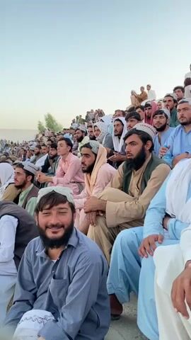 A medium shot captures a crowd of men seated on bleachers, their faces turned towards an unseen event. The men are predominantly wearing traditional Islamic attire, including shalwar kameez and turbans or skullcaps, in various shades of white, blue, and earth tones. The man in the foreground on the left is wearing a grey shirt and a white skullcap, smiling at the camera. The background reveals more spectators stretching up the incline of the bleachers, against a backdrop of a pale, hazy sky and a hint of distant greenery. The lighting suggests it is either early morning or late afternoon. The overall atmosphere appears to be one of communal gathering and observation.