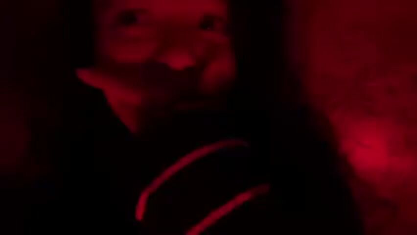 In a darkened setting with red lighting, a young individual with dark hair is visible from the chest up. The person appears to be listening to music. The red lighting casts strong shadows, making it difficult to discern fine details. The background appears to have a floral pattern. The individual's hand is near their face.