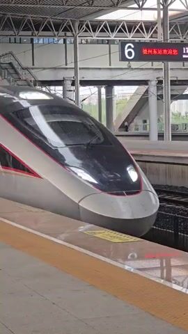 A high-speed train, predominantly white with red and gold accents, is shown at a station platform. The train has a sleek, aerodynamic design. A digital display above the train indicates track number 6 and the destination Dezhou East Station, with a departure time of 17:22. The platform has a yellow textured safety line along its edge. Reflections of people and the station roof are visible on the train's windows. A woman with long, dark, wavy hair, wearing a black top, is partially visible walking away from the camera on the platform. The train appears to be stationary at the platform.